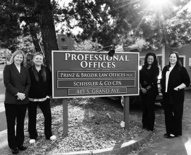Black-and-white group photo of four Prinz & Brozik Law Offices team members standing beside their office sign at 445 S. Grand Avenue.
